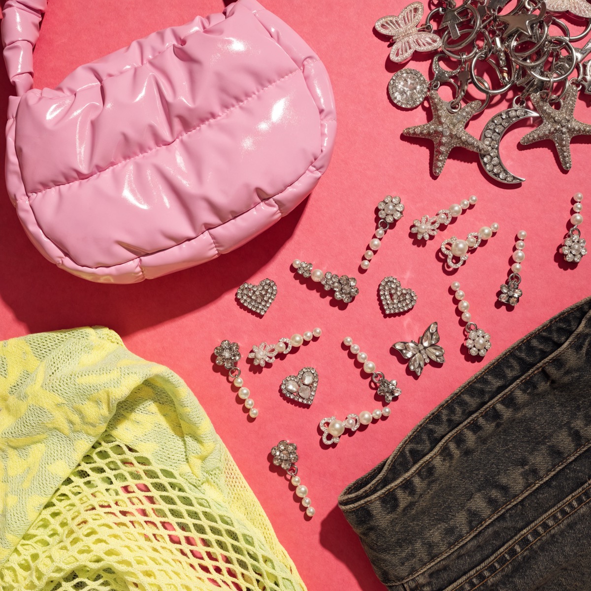 A high-saturation Y2K flat lay on rose paper: a glossy pink puffer mini bag, charm keychains, a mesh top, rhinestone clips and folded denim.