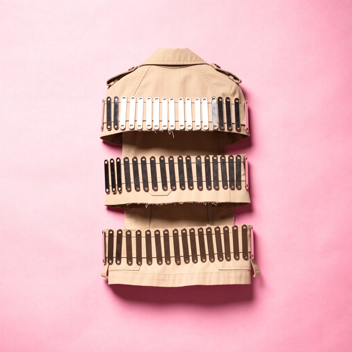 A khaki trench coat laid flat on pink paper, cut into three horizontal sections and pinned back together with dense rows of safety pins.