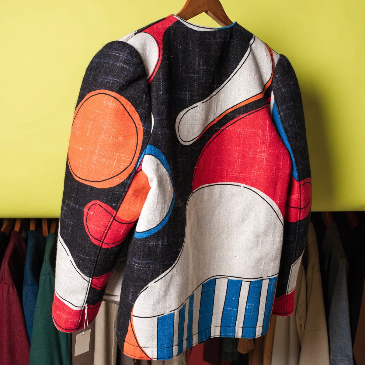 An archival 1980s Tokyo designer jacket with oversized shoulders and a bold graphic print, on a hanger against a chartreuse wall, a blank resale tag at the cuff.